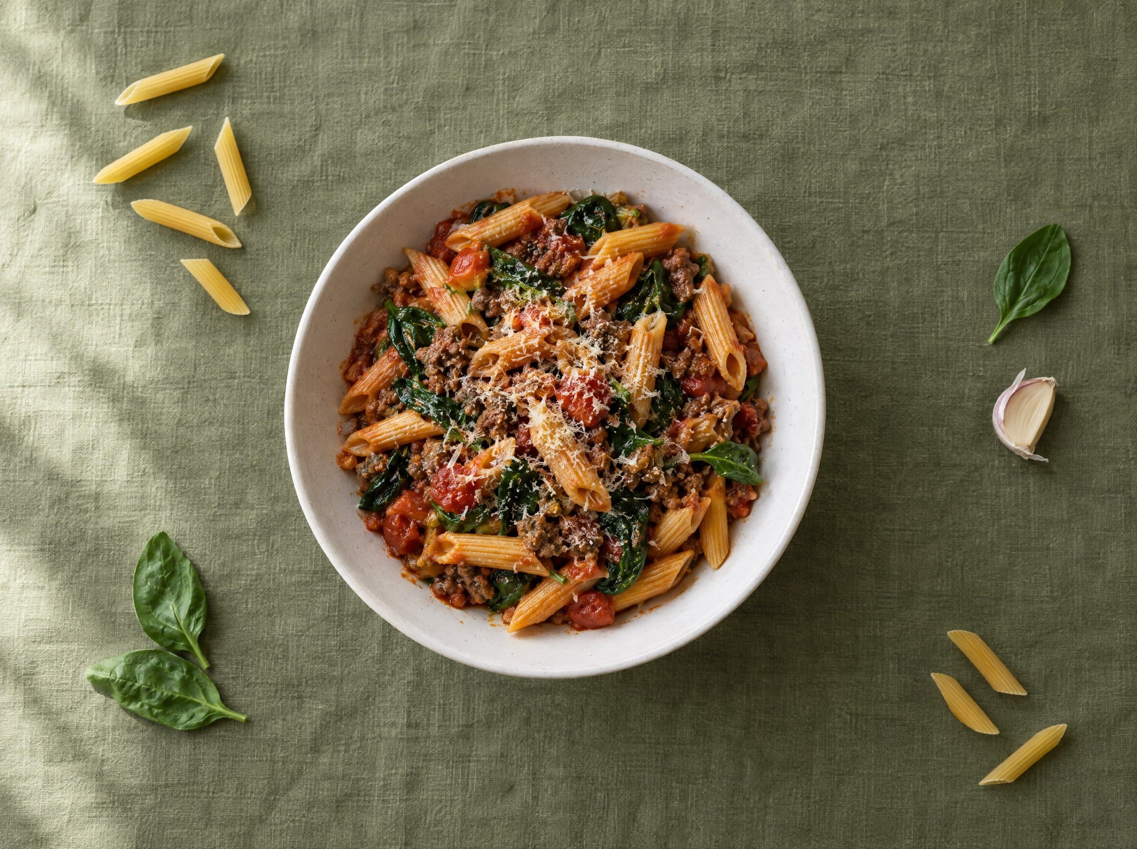 Penne met tomatensaus, gehakt, spinazie en geraspte kaas in een witte kom op een olijfgroen linnen kleed